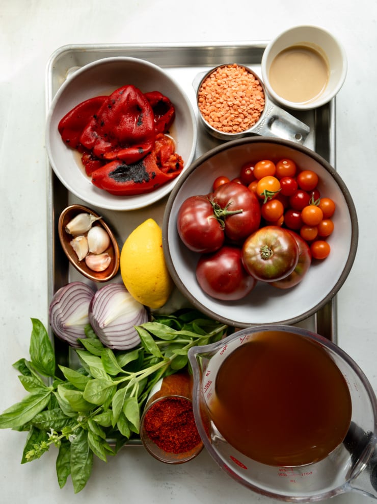 Overhead flat-lay of all ingredients for red lentil tomato soup arranged on a metal baking tray: whole roasted red peppers, a measuring cup of dried red lentils, a small cup of tahini, whole heirloom and cherry tomatoes in a bowl, garlic cloves, a whole lemon, halved red onion, a bunch of fresh basil, a small dish of smoked paprika or chili powder, and a large measuring cup of vegetable broth.
