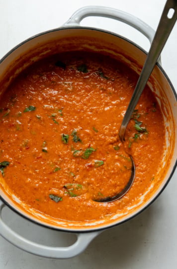 Overhead view of red lentil tomato soup in a white enameled Dutch oven, still in the pot before serving. The soup has a thick, creamy texture with flecks of chopped fresh basil stirred throughout, and a silver ladle rests inside, handle propped against the pot's rim.