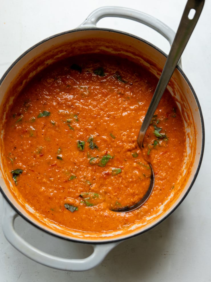 Overhead view of red lentil tomato soup in a white enameled Dutch oven, still in the pot before serving. The soup has a thick, creamy texture with flecks of chopped fresh basil stirred throughout, and a silver ladle rests inside, handle propped against the pot's rim.