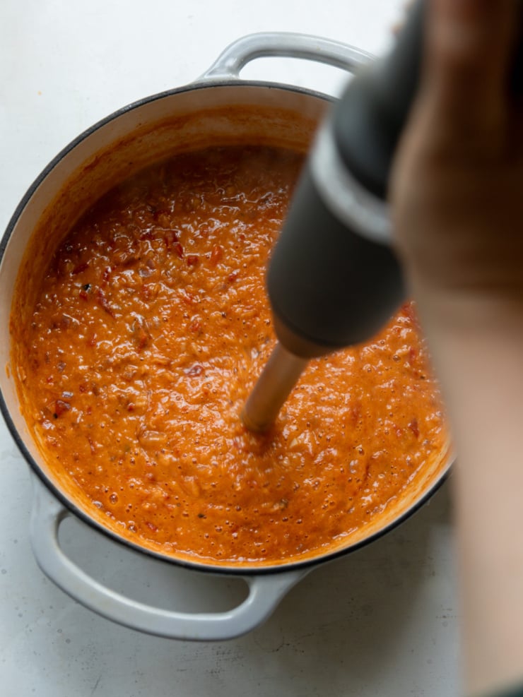 Overhead action shot of an immersion blender pureeing red lentil tomato soup directly in a white Dutch oven. The blender head is submerged in the chunky orange-red mixture, mid-blend, with visible bits of tomato, roasted red pepper, and lentils not yet fully smooth; a person's arm is partially visible blurred in the foreground.