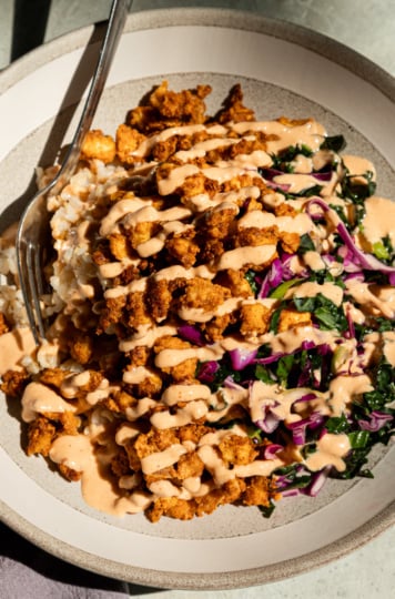 Overhead shot of a crispy tofu bowl with golden popcorn tofu piled over brown rice, drizzled generously with spicy garlic tahini sauce, and served alongside a shredded kale and red cabbage slaw, fork resting on the side of the bowl.