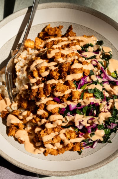 Overhead shot of a crispy tofu bowl with golden popcorn tofu piled over brown rice, drizzled generously with spicy garlic tahini sauce, and served alongside a shredded kale and red cabbage slaw, fork resting on the side of the bowl.