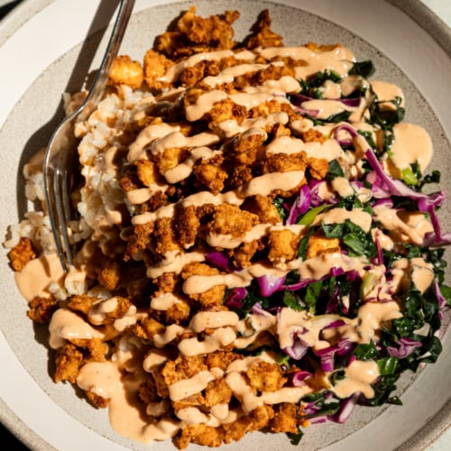 Overhead shot of a crispy tofu bowl with golden popcorn tofu piled over brown rice, drizzled generously with spicy garlic tahini sauce, and served alongside a shredded kale and red cabbage slaw, fork resting on the side of the bowl.