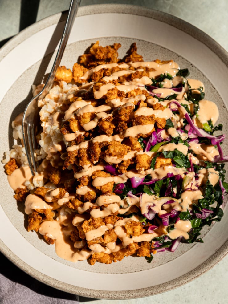 Overhead shot of a crispy tofu bowl with golden popcorn tofu piled over brown rice, drizzled generously with spicy garlic tahini sauce, and served alongside a shredded kale and red cabbage slaw, fork resting on the side of the bowl.