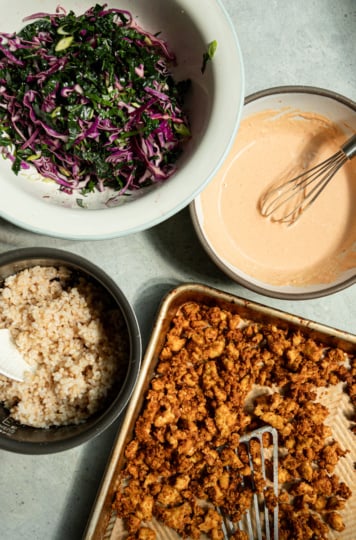 Four components of a crispy tofu bowl recipe arranged for assembly: a bowl of massaged kale and red cabbage slaw, a bowl of creamy orange-hued spicy garlic tahini sauce with a whisk, a container of cooked brown rice, and a sheet pan of freshly baked crispy popcorn tofu pieces.
