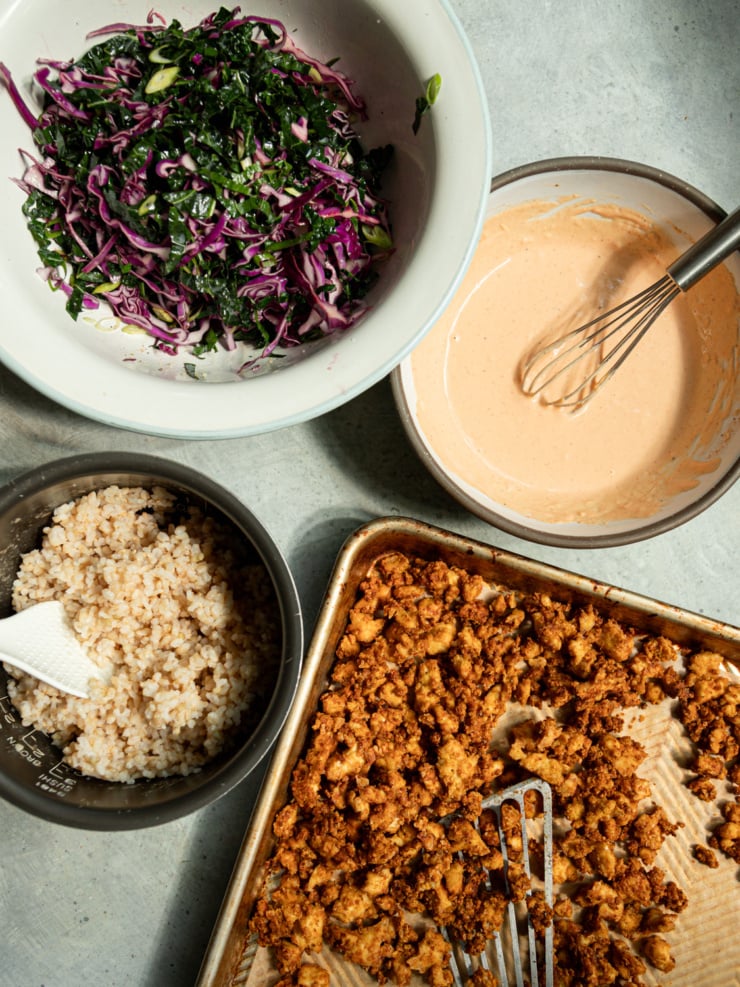 Four components of a crispy tofu bowl recipe arranged for assembly: a bowl of massaged kale and red cabbage slaw, a bowl of creamy orange-hued spicy garlic tahini sauce with a whisk, a container of cooked brown rice, and a sheet pan of freshly baked crispy popcorn tofu pieces.