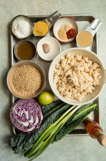 Overhead flatlay of ingredients for crispy popcorn tofu bowls, including crumbled tofu, brown rice, half a head of red cabbage, kale leaves, green onions, a lime, hot sauce, olive oil, a garlic clove, nutritional yeast, arrowroot starch, tahini, and a spice bowl of garlic powder, onion powder, and paprika, all arranged on a metal sheet pan.