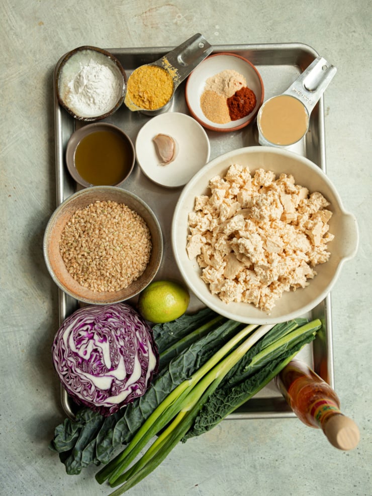 Overhead flatlay of ingredients for crispy popcorn tofu bowls, including crumbled tofu, brown rice, half a head of red cabbage, kale leaves, green onions, a lime, hot sauce, olive oil, a garlic clove, nutritional yeast, arrowroot starch, tahini, and a spice bowl of garlic powder, onion powder, and paprika, all arranged on a metal sheet pan.