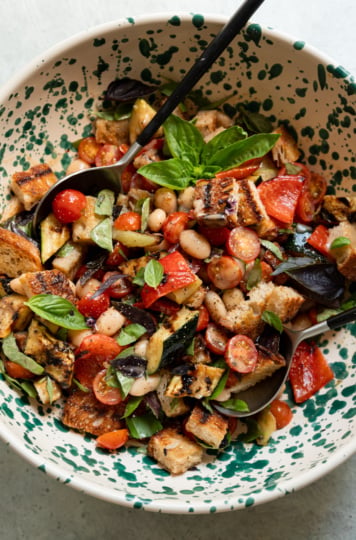 Overhead view of a speckled ceramic bowl filled with grilled vegetable panzanella salad, tossed and garnished with a basil sprig, with two serving spoons resting in the bowl.