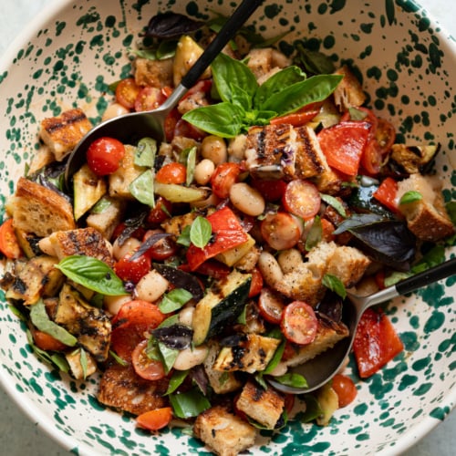 Overhead view of a speckled ceramic bowl filled with grilled vegetable panzanella salad, tossed and garnished with a basil sprig, with two serving spoons resting in the bowl.