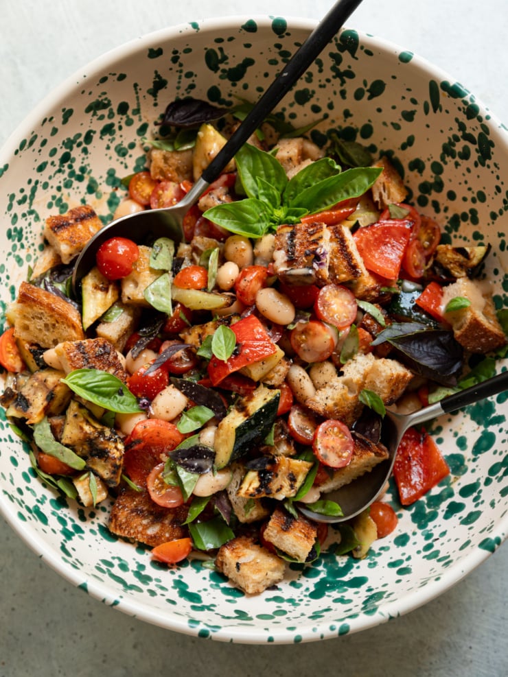Overhead view of a speckled ceramic bowl filled with grilled vegetable panzanella salad, tossed and garnished with a basil sprig, with two serving spoons resting in the bowl.