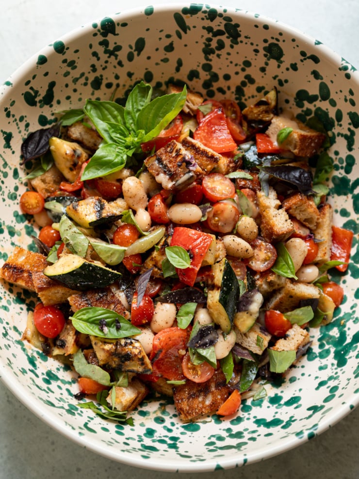 Overhead close-up of the panzanella salad in a speckled ceramic bowl, showing grilled zucchini, croutons, cherry tomatoes, cannellini beans, and basil, garnished with a basil sprig on top.