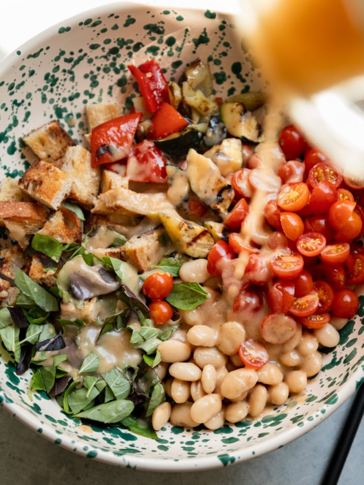 Overhead view of dressing being poured over the panzanella salad ingredients in a speckled ceramic bowl before tossing.