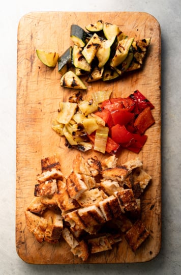 Overhead view of a wooden cutting board with grilled zucchini, grilled red bell pepper and banana pepper pieces, and cubed grilled sourdough bread arranged in separate piles.