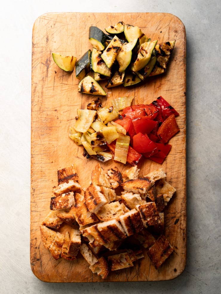 Overhead view of a wooden cutting board with grilled zucchini, grilled red bell pepper and banana pepper pieces, and cubed grilled sourdough bread arranged in separate piles.