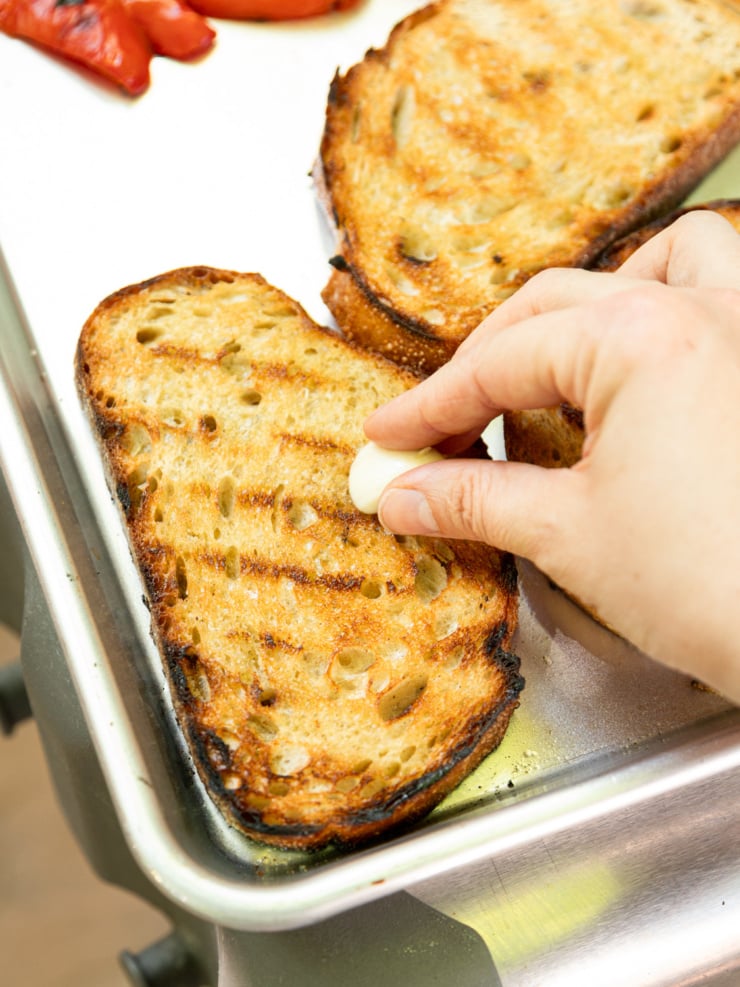 Close-up of a hand rubbing a halved garlic clove over grilled sourdough bread slices on a sheet pan, with grilled red pepper visible in the background.