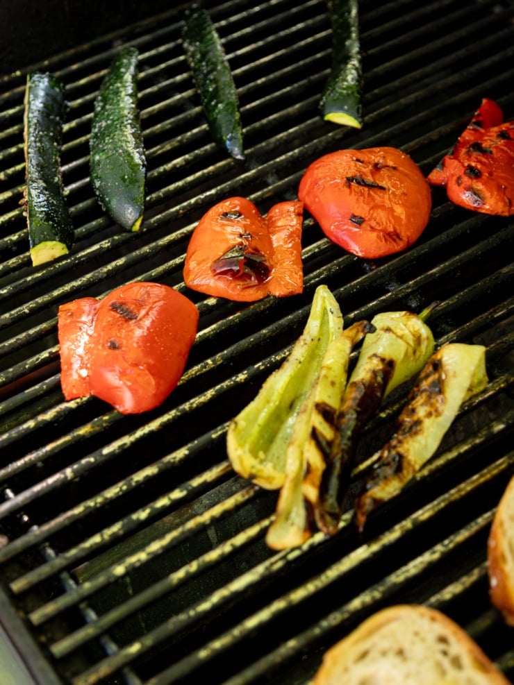 Close-up overhead view of grilled zucchini spears, red bell pepper halves, and banana peppers with char marks on a outdoor grill grate.
