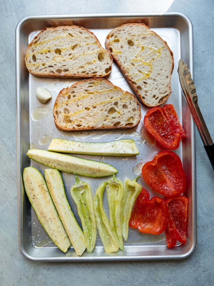 Overhead view of a sheet pan with three slices of olive oil-brushed grilled sourdough bread, a halved garlic clove, and raw seasoned zucchini spears, banana peppers, and red bell pepper pieces ready for grilling, with tongs resting on the side.