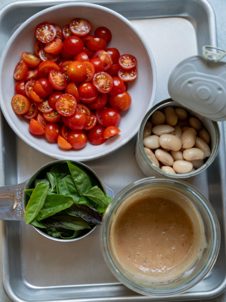 Overhead view of halved cherry tomatoes in a bowl, a measuring cup of fresh basil leaves, an open can of cannellini beans, and a jar of dressing, arranged on a sheet pan.