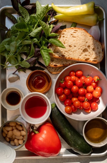 Overhead view of a sheet pan with ingredients for the salad laid out for prep: fresh basil, sourdough bread slices, banana peppers, a shallot, small bowls of red wine vinegar and vegan Worcestershire sauce, a bowl of cherry tomatoes, a can of cannellini beans, a red bell pepper, a zucchini, and a small bowl of olive oil.