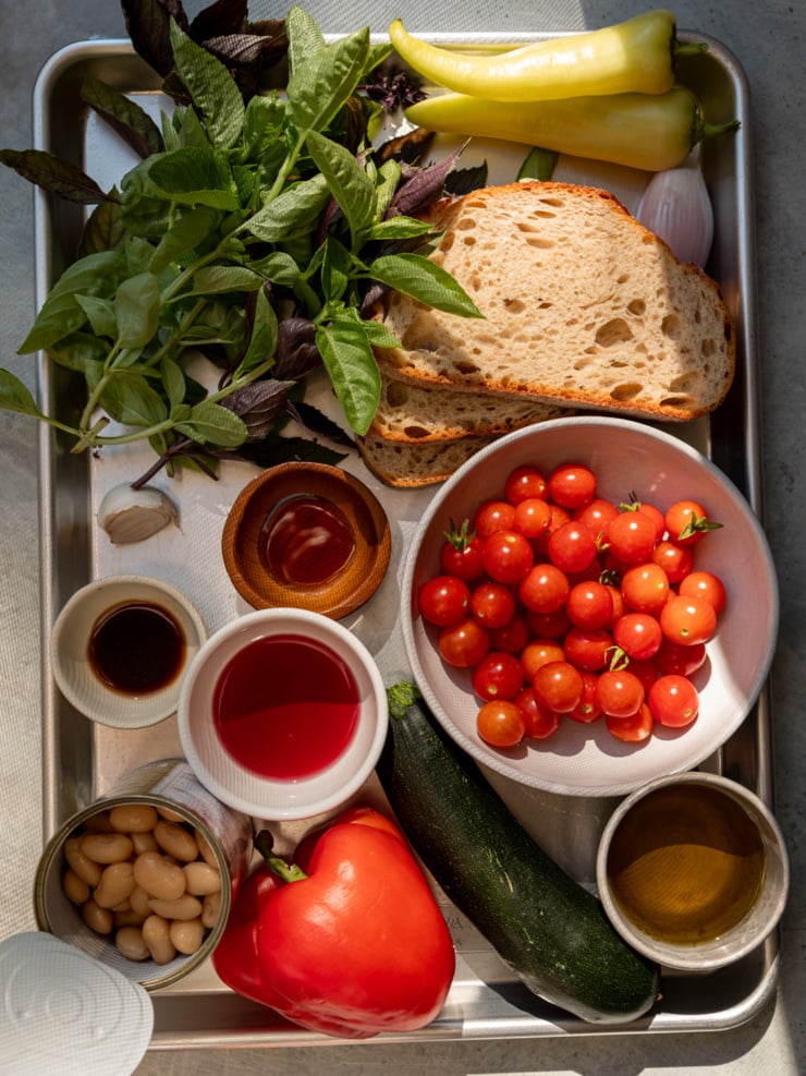 Overhead view of a sheet pan with ingredients for the salad laid out for prep: fresh basil, sourdough bread slices, banana peppers, a shallot, small bowls of red wine vinegar and vegan Worcestershire sauce, a bowl of cherry tomatoes, a can of cannellini beans, a red bell pepper, a zucchini, and a small bowl of olive oil.