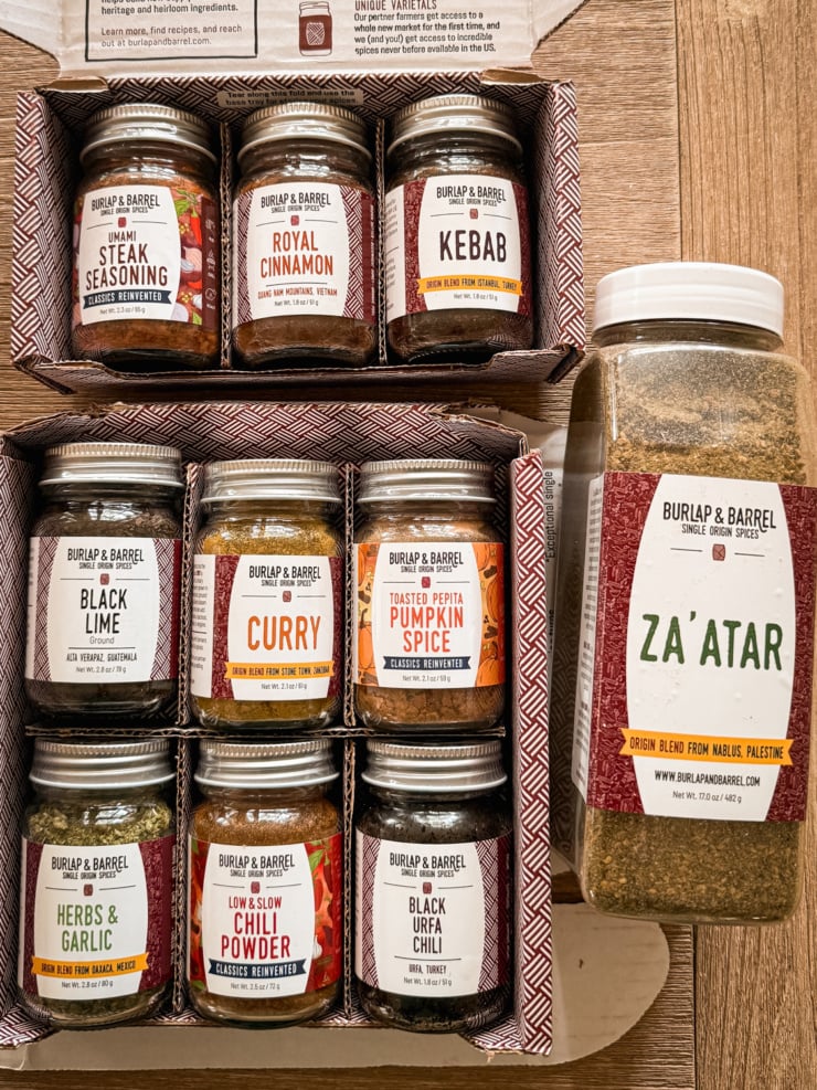 Two open boxes of Burlap & Barrel spice jars, including Umami Steak Seasoning, Royal Cinnamon, Kebab, Black Lime, Curry, Toasted Pepita Pumpkin Spice, Herbs & Garlic, Low & Slow Chili Powder, and Black Urfa Chili, next to a large jar of Za'atar seasoning.