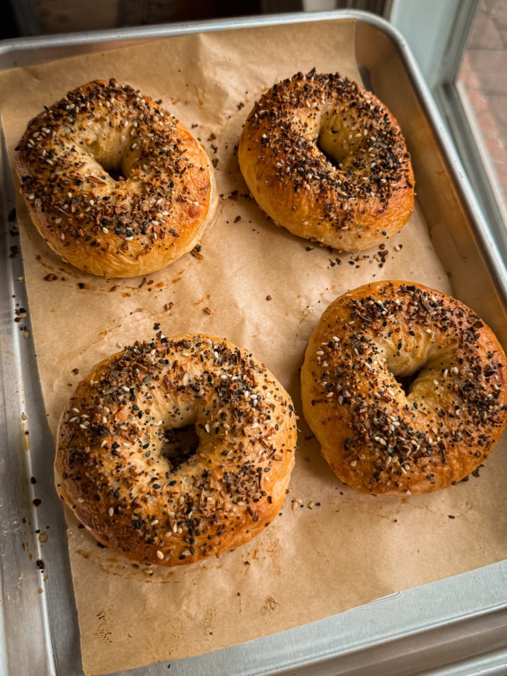 Four freshly baked everything bagels topped with sesame seeds, poppy seeds, and dried garlic and onion, resting on parchment paper in a metal baking tray.