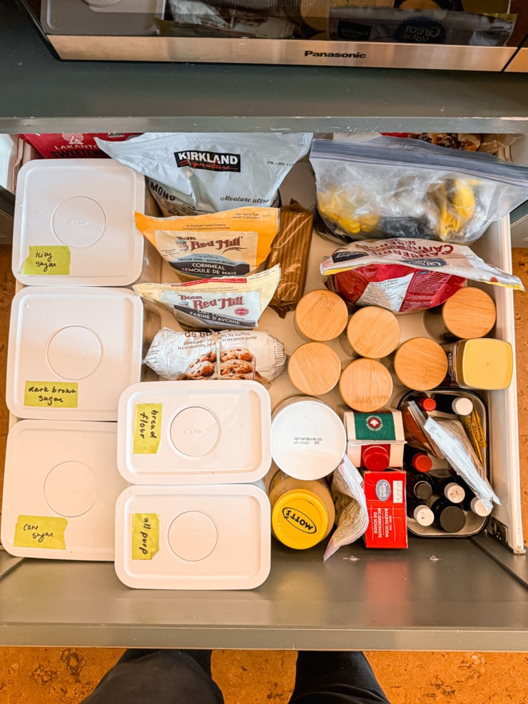 Overhead view of an organized kitchen drawer with labeled OXO containers (icing sugar, dark brown sugar, cane sugar, bread flour, all-purpose flour) alongside cornmeal, oat flour, and small jars with wooden lids.