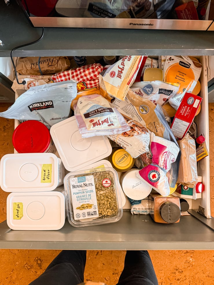 Overhead view of a cluttered, overflowing kitchen drawer packed with flour bags, baking powder, pumpkin seeds, and mismatched containers.