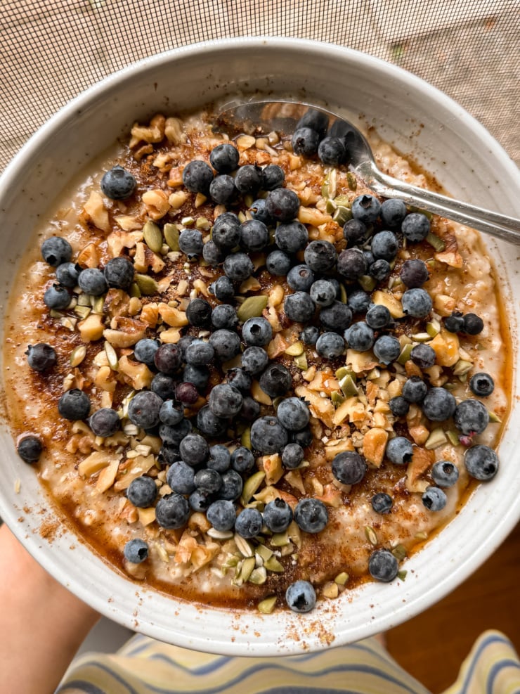 Overhead view of a bowl of oatmeal topped with a generous pile of fresh blueberries, chopped walnuts, pumpkin seeds, ground flax, cinnamon and maple syrup, with a spoon resting in the bowl.