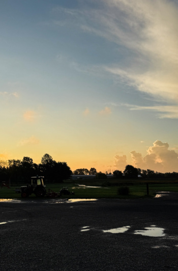 A pastel sunrise over an open field, with a silhouetted tractor and tree line against a glowing orange horizon, wispy clouds streaked across a blue sky, and rain puddles reflecting the light in the foreground pavement.