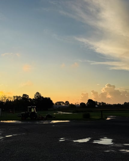 A pastel sunrise over an open field, with a silhouetted tractor and tree line against a glowing orange horizon, wispy clouds streaked across a blue sky, and rain puddles reflecting the light in the foreground pavement.