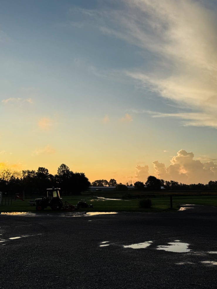 A pastel sunrise over an open field, with a silhouetted tractor and tree line against a glowing orange horizon, wispy clouds streaked across a blue sky, and rain puddles reflecting the light in the foreground pavement.