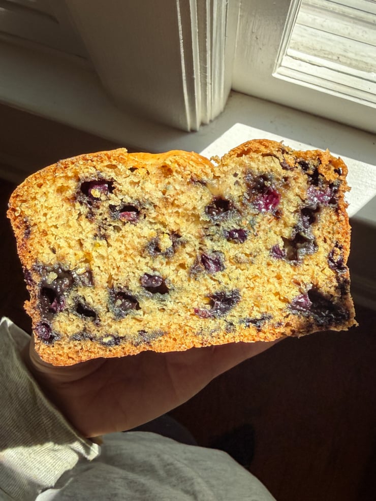 A hand holding up a thick slice of blueberry quick bread in a sunlit window, the golden crumb studded with juicy blueberries.