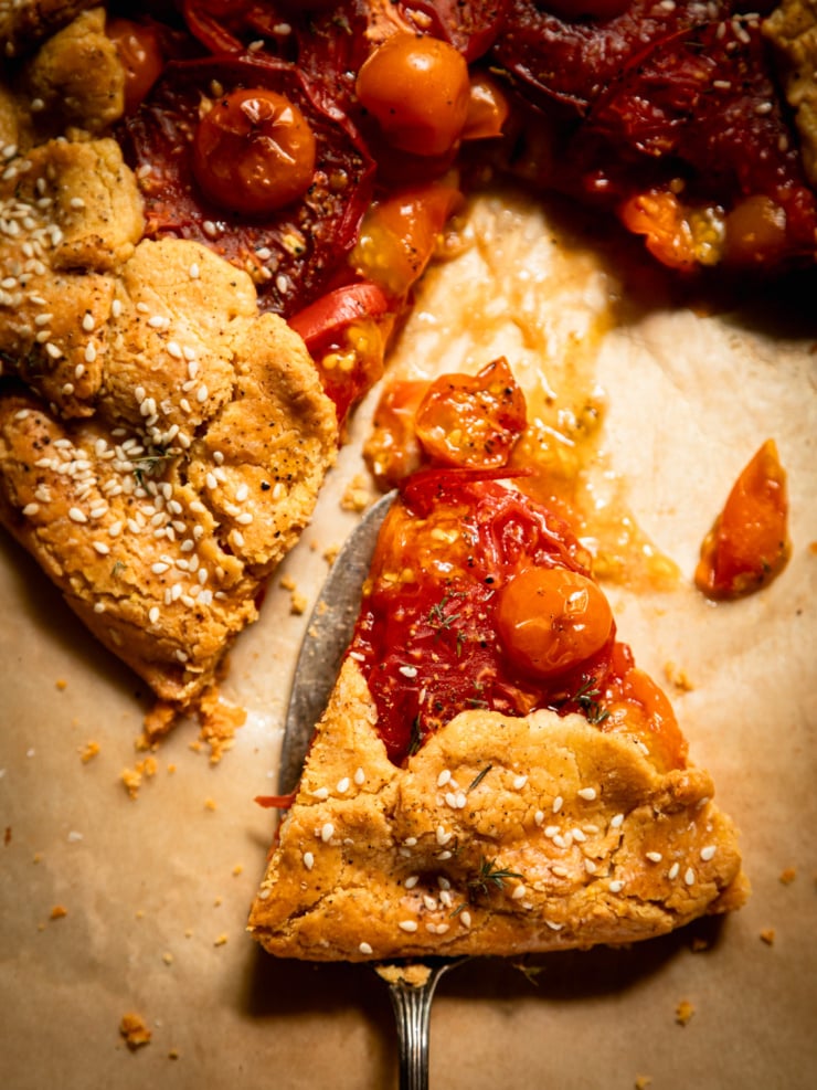 Overhead close-up of a slice of tomato galette being lifted with a knife, showing the gluten-free chickpea crust edge studded with sesame seeds beside glossy roasted tomatoes and cherry tomatoes on parchment paper.