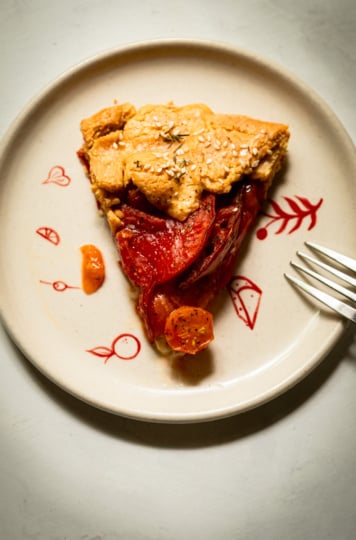 Overhead shot of a single slice of tomato galette on a cream plate with hand-painted red botanical designs, showing layered tomato slices and a cherry tomato beneath a sesame-topped crust, with a fork resting on the right side.