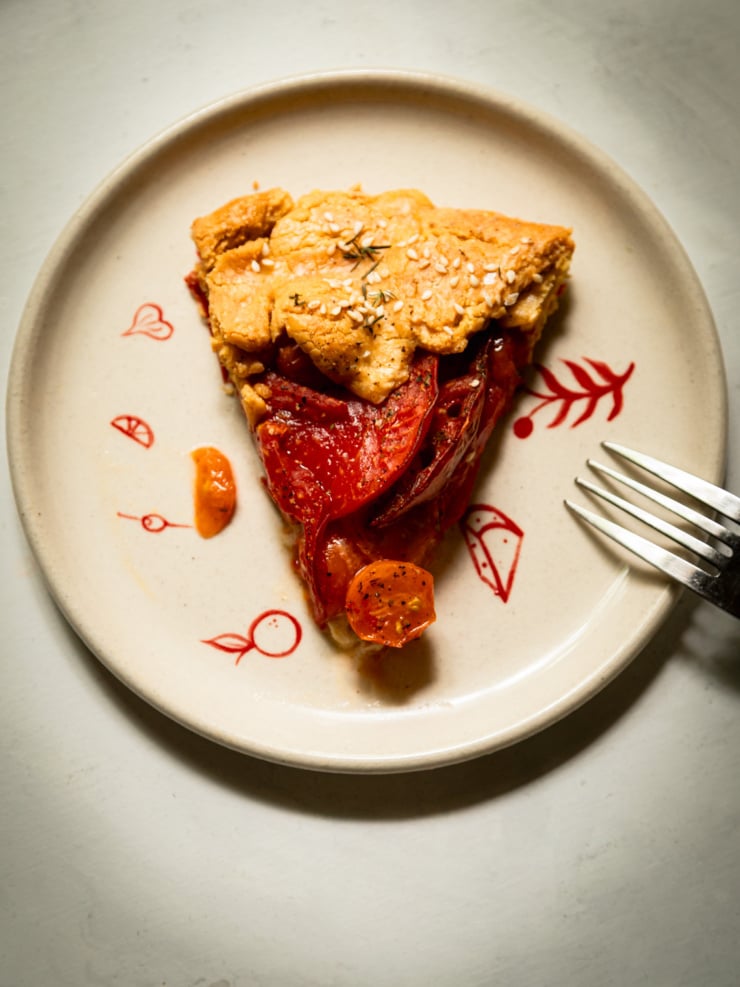 Overhead shot of a single slice of tomato galette on a cream plate with hand-painted red botanical designs, showing layered tomato slices and a cherry tomato beneath a sesame-topped crust, with a fork resting on the right side.