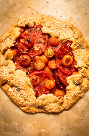 Overhead view of a whole baked tomato galette on parchment paper, its golden chickpea-cornmeal crust folded over the edges and topped with sesame seeds, framing a center of overlapping red and orange tomato slices and cherry tomatoes.