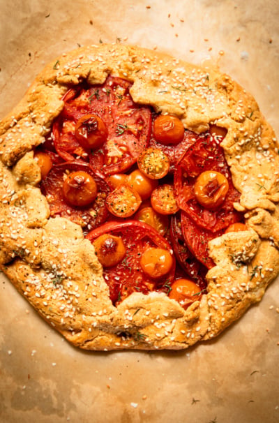 Overhead view of a whole baked tomato galette on parchment paper, its golden chickpea-cornmeal crust folded over the edges and topped with sesame seeds, framing a center of overlapping red and orange tomato slices and cherry tomatoes.