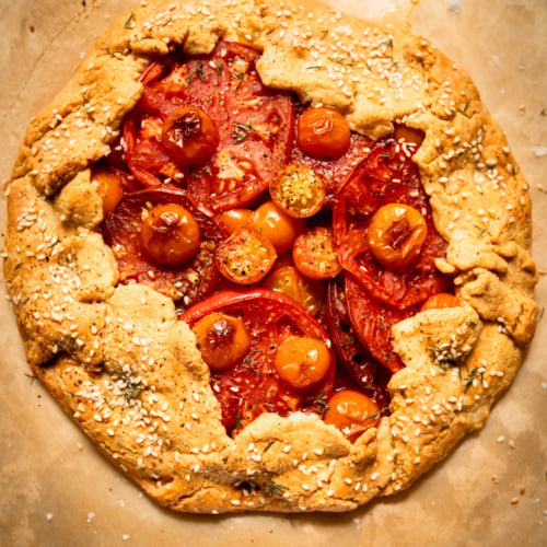 Overhead view of a whole baked tomato galette on parchment paper, its golden chickpea-cornmeal crust folded over the edges and topped with sesame seeds, framing a center of overlapping red and orange tomato slices and cherry tomatoes.