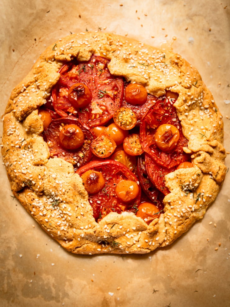 Overhead view of a whole baked tomato galette on parchment paper, its golden chickpea-cornmeal crust folded over the edges and topped with sesame seeds, framing a center of overlapping red and orange tomato slices and cherry tomatoes.