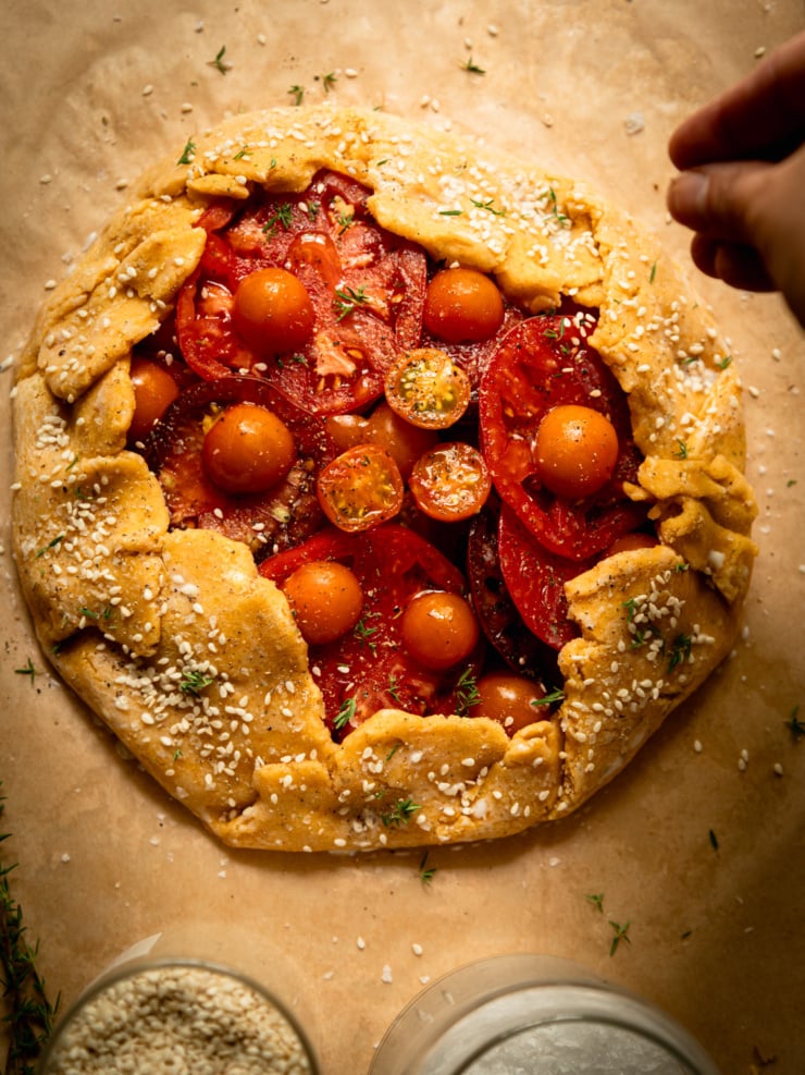Overhead view of the whole, unbaked and assembled tomato galette with a hand reaching in from the upper right to sprinkle sesame seeds over the chickpea pastry.