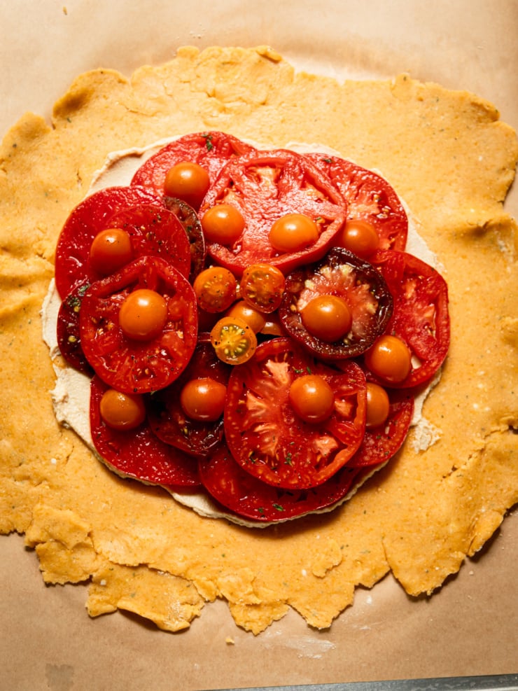 Overhead shot of unbaked galette dough rolled out on parchment paper, with sliced heirloom tomatoes and halved cherry tomatoes mounded in the center over a layer of soft vegan cheese, ready to be folded.