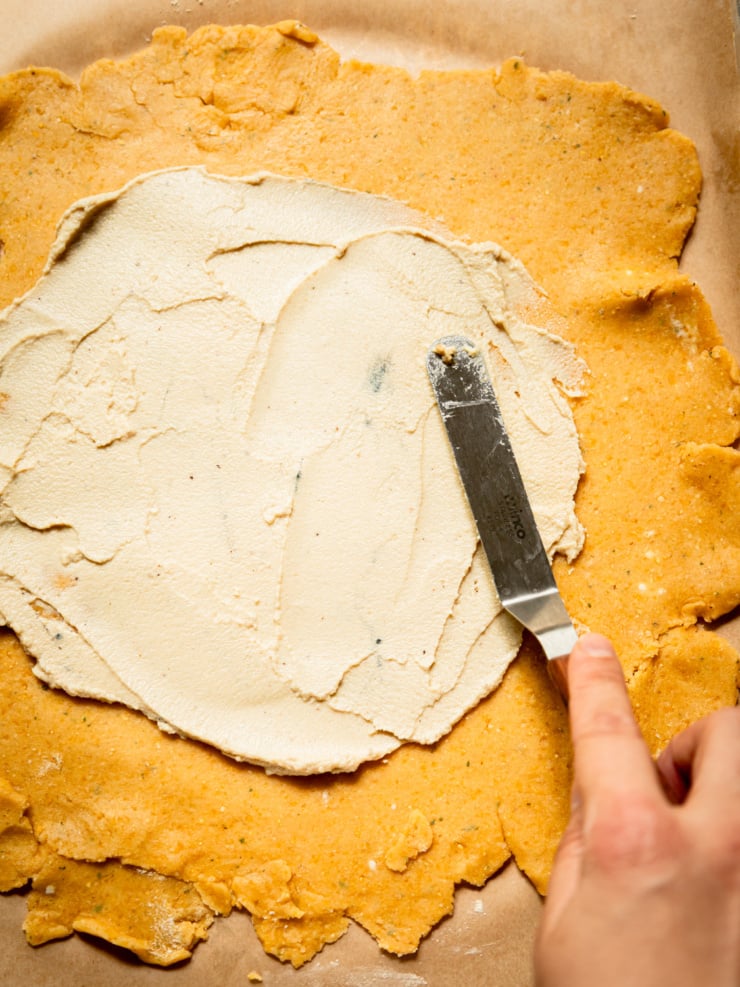 Close-up overhead shot of a small offset spatula spreading soft vegan cheese in a circle over rolled-out golden chickpea-cornmeal galette dough.