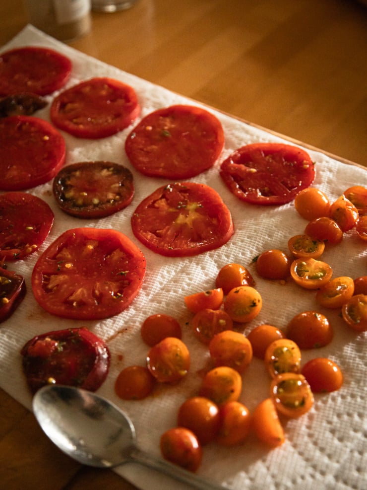 Slight overhead shot of sliced red and heirloom tomatoes and halved orange cherry tomatoes laid out on paper towels to drain, with a spoon resting at the lower left.