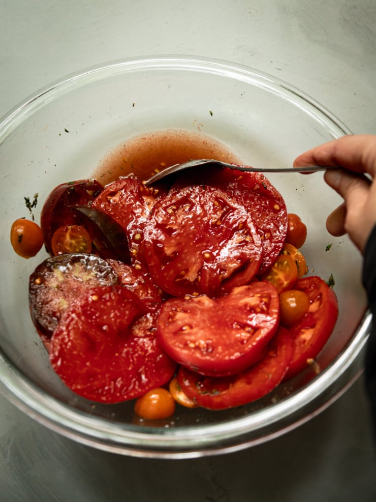 Overhead shot of a hand using a spoon to toss sliced tomatoes and cherry tomatoes with seasoning in a clear glass bowl.