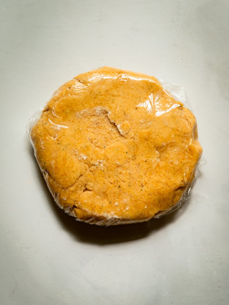 Overhead shot of a disc of golden chickpea galette dough wrapped in plastic wrap, resting on a white surface.