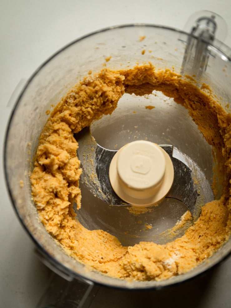 Overhead close-up inside a food processor bowl showing golden chickpea galette dough clumped around the blade after mixing.