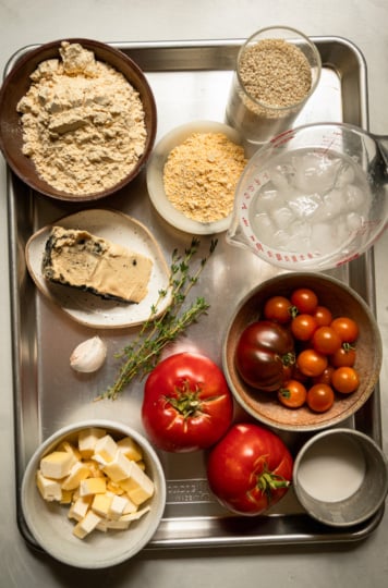 Overhead shot of galette ingredients arranged on a metal tray: chickpea flour, cornmeal, sesame seeds, a cup of ice water, a wedge of soft vegan cheese, fresh thyme sprigs, a garlic clove, cubed vegan butter, two whole tomatoes, a bowl of cherry tomatoes, and a small cup of plant milk.