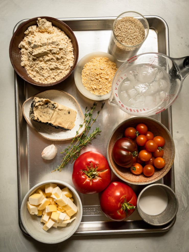 Overhead shot of galette ingredients arranged on a metal tray: chickpea flour, cornmeal, sesame seeds, a cup of ice water, a wedge of soft vegan cheese, fresh thyme sprigs, a garlic clove, cubed vegan butter, two whole tomatoes, a bowl of cherry tomatoes, and a small cup of plant milk.